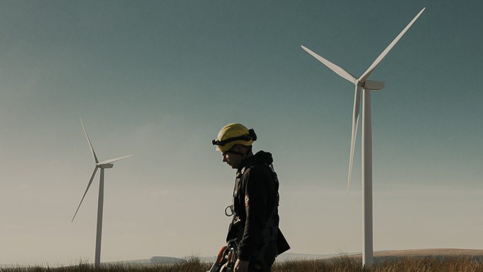 A person wearing a yellow hardhat, standing in front of two wind turbines